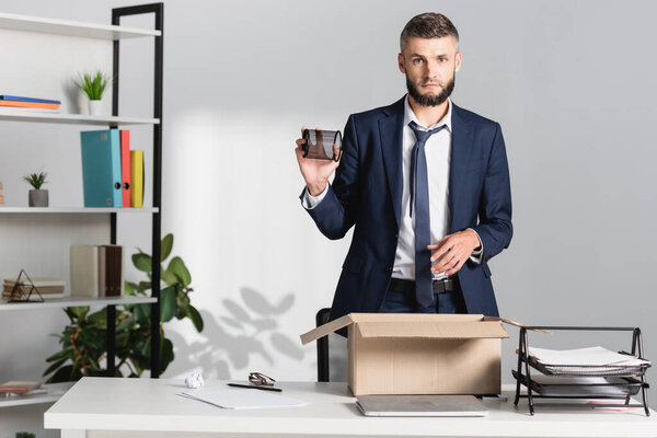 Fired businessman holding stationery near carton box, laptop and papers on table 