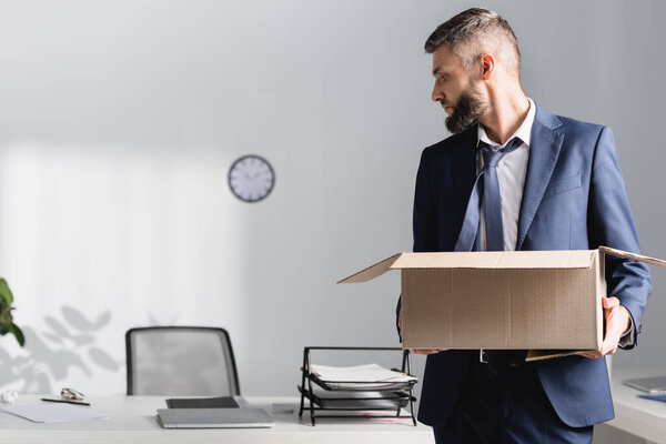 Fired businessman holding carton box near office table on blurred background in office 