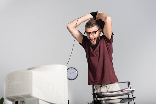 Screaming businessman holding keyboard near computer monitor and papers on blurred foreground in office 