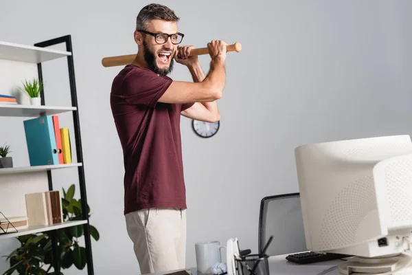 拿着棒球棒站在电脑前尖叫的商人在模糊的前景上 — 图库照片