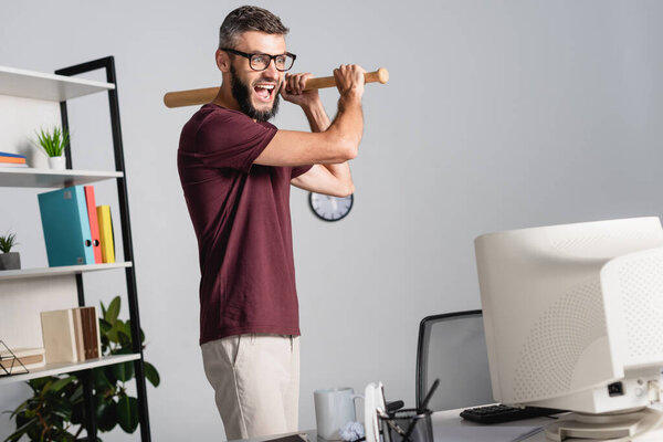 Screaming businessman with baseball bat standing near computer on blurred foreground 