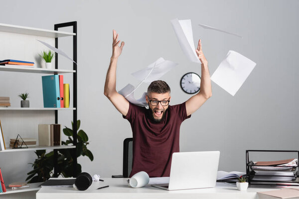 Stressed businessman screaming while throwing papers near laptop on office table 