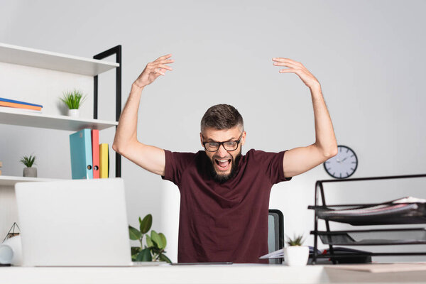 Angry businessman screaming near laptop and stationery on blurred foreground in office 