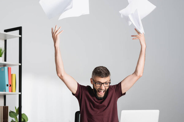 Tensed and screaming businessman throwing documents near laptop on blurred foreground in office 