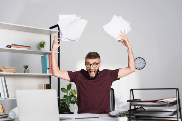 Screaming businessman throwing papers near laptop on blurred foreground in office 