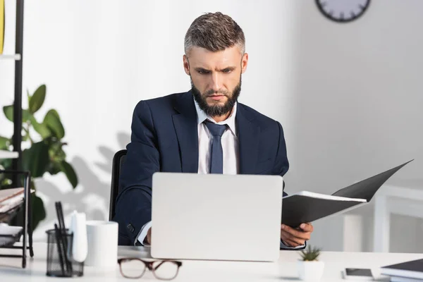 Geconcentreerde Zakenman Houdt Papieren Map Buurt Van Laptop Wazig Voorgrond — Stockfoto