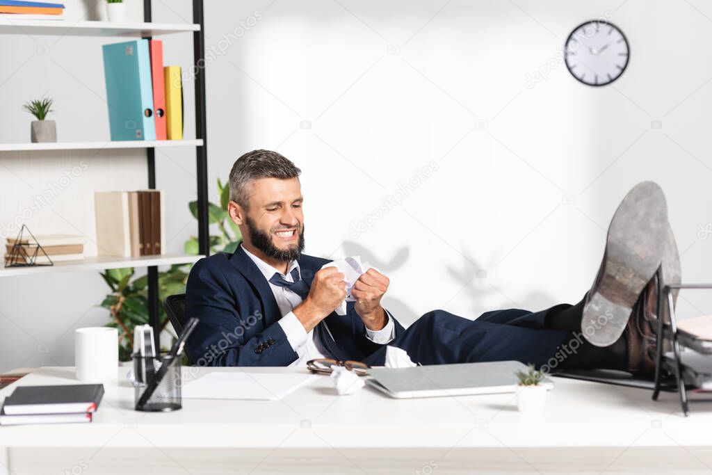 Stressed businessman holding clumped paper near laptop and stationery on blurred foreground 