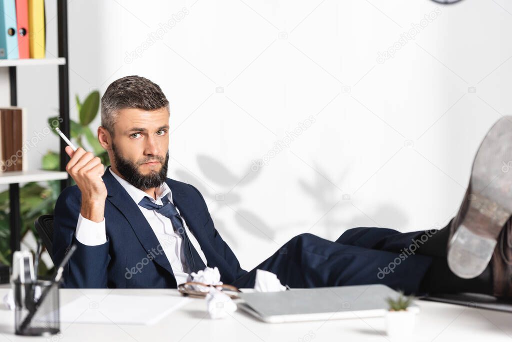 Businessman holding pencil near laptop and clumped paper on table on blurred foreground