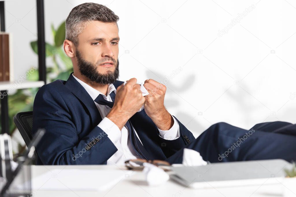 Stressed businessman holding clumped paper near laptop and stationery on blurred foreground in office 
