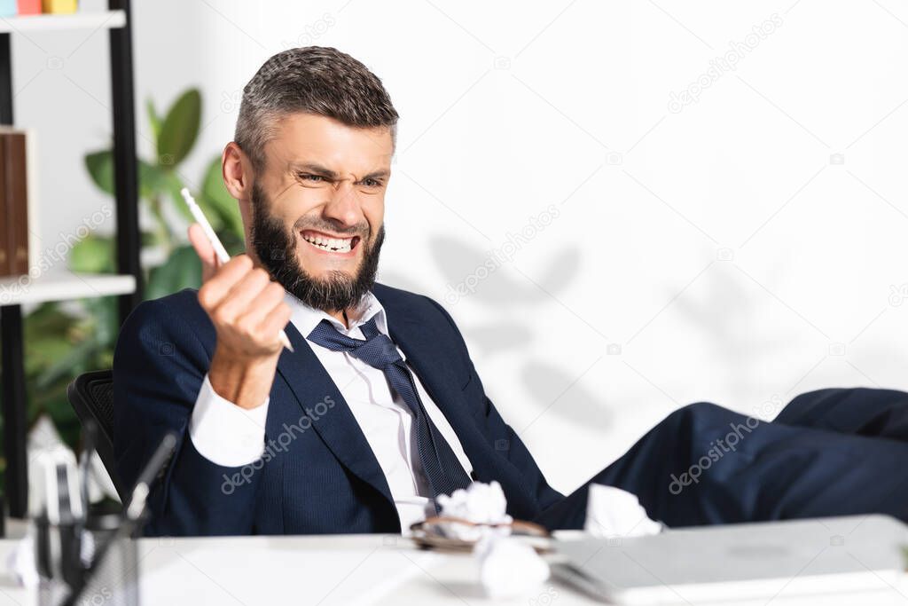 Nervous businessman holding pencil near clumped paper and laptop on blurred foreground in office 