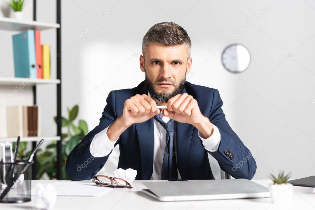 Businessman holding pencil near laptop and clumped paper on blurred foreground in office 