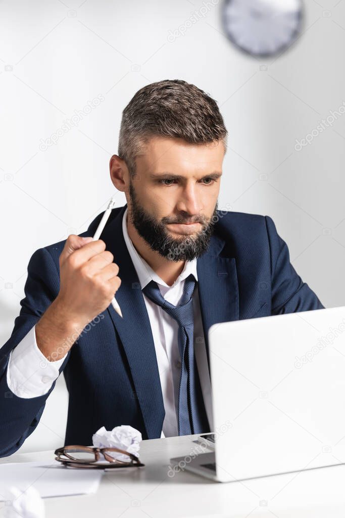 Stressed businessman holding broken pencil near gadgets, clumped paper and eyeglasses on blurred foreground in office 
