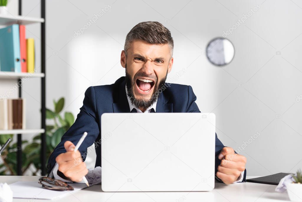 Angry businessman screaming while holding broken pencil near laptop on blurred foreground in office 