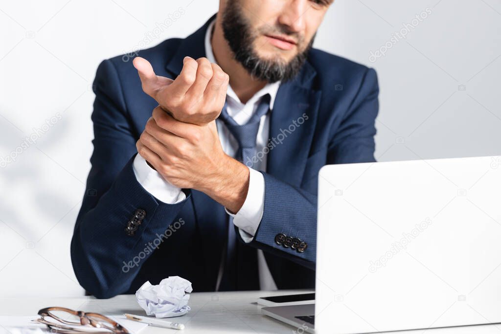 Cropped view of businessman feeling arm pain near gadgets and clumped paper on table 