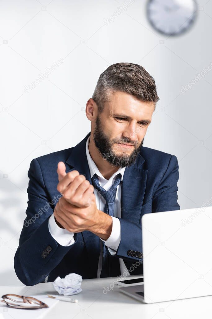 Businessman touching arm while suffering from pain near gadgets on working table on blurred foreground in office 