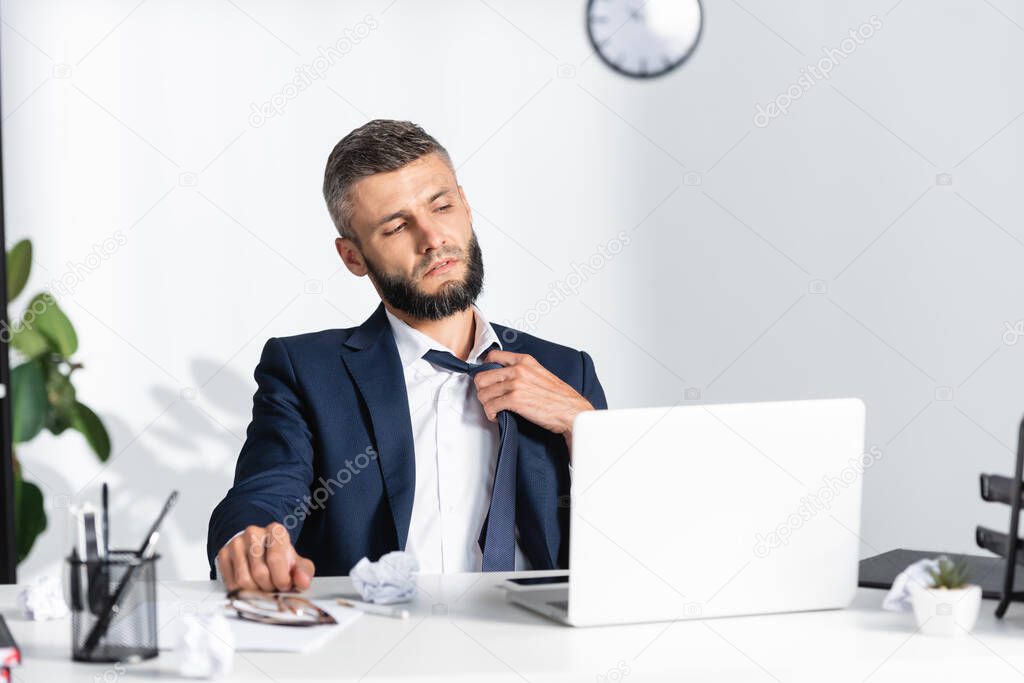 Businessman touching tie while suffering from heat near gadgets and stationery on blurred foreground 