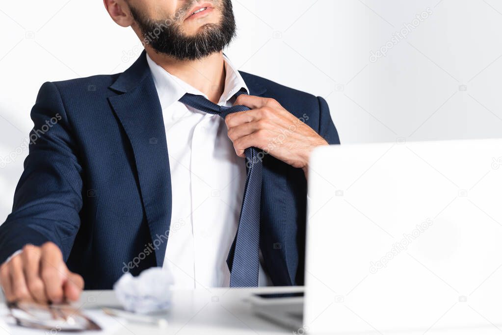 Cropped view of businessman touching tie while feeling hot near laptop on blurred foreground 