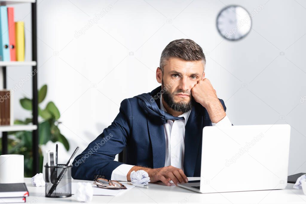 Pensive businessman looking at camera near laptop, stationery and clumped paper on blurred foreground in office 