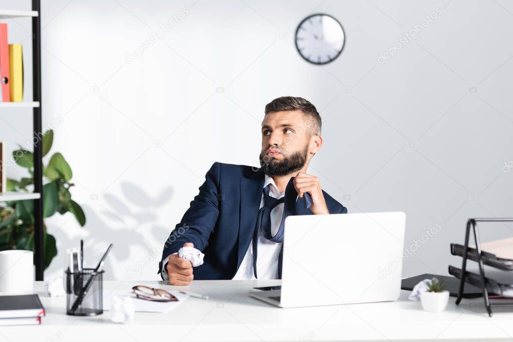Pensive businessman touching tie and holding clumped paper near laptop and stationery on blurred foreground in office 