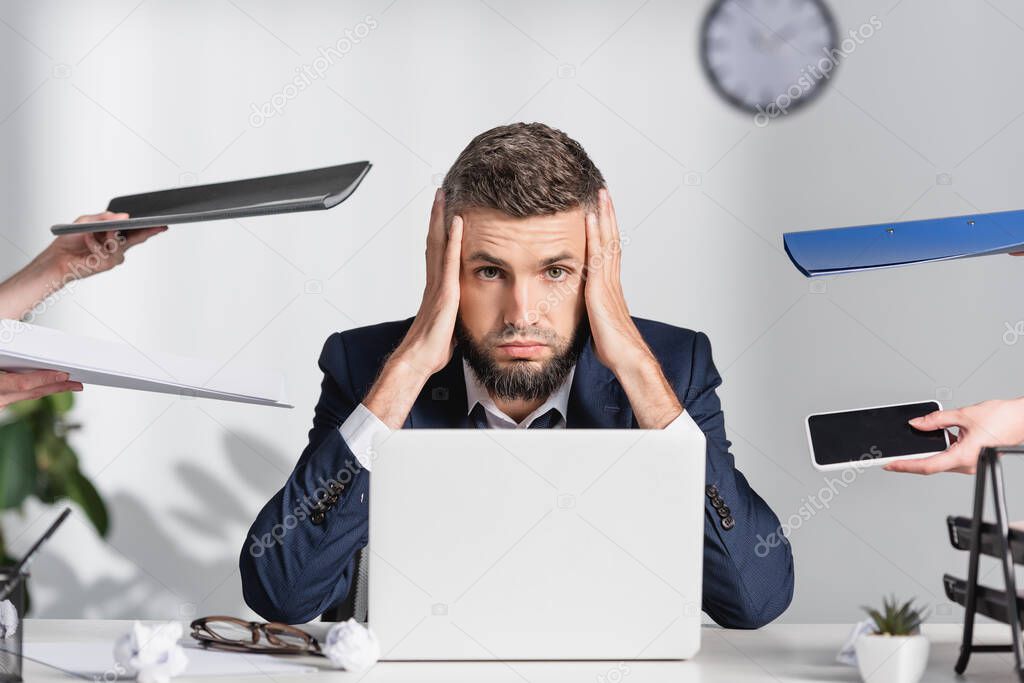 Tired businessman looking at camera near colleagues with paper folders and smartphone in office 