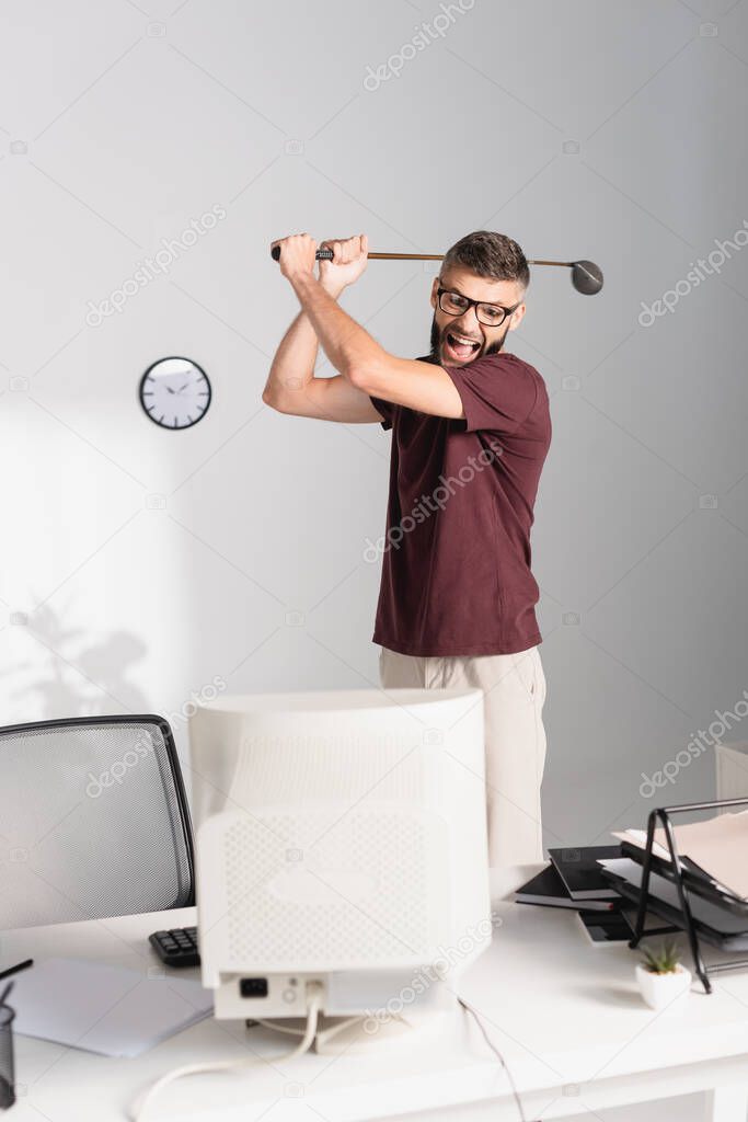 Stressed businessman holding golf club near computer and papers on blurred foreground in office 