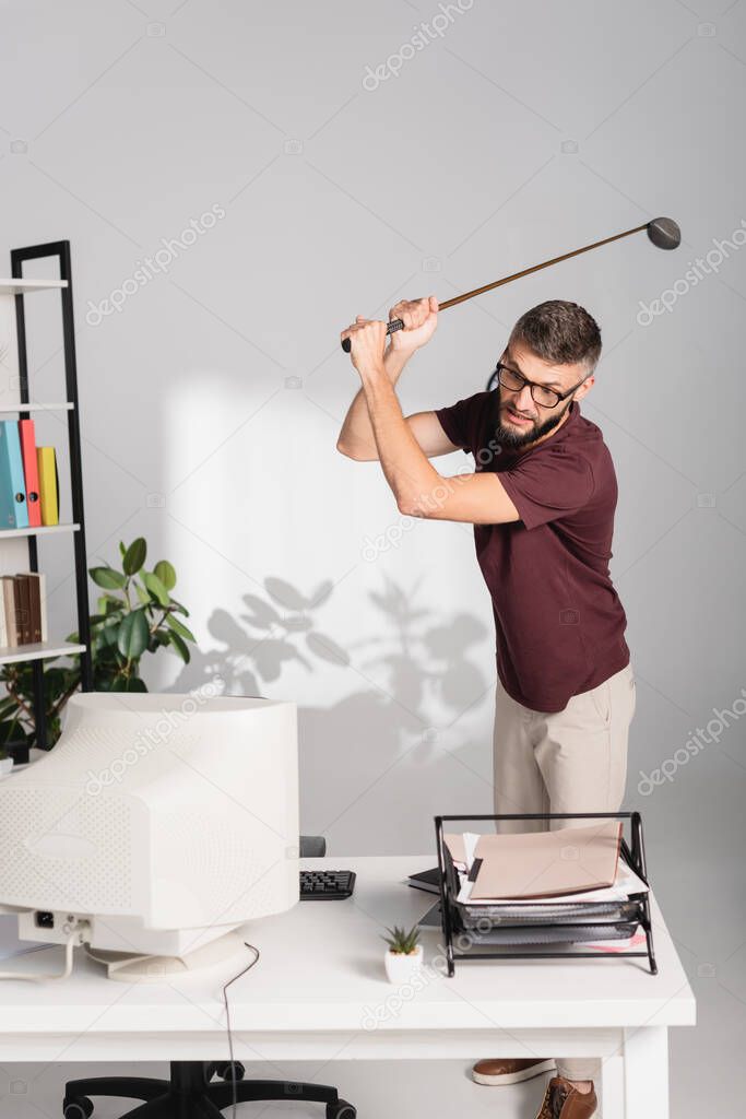 Angry businessman with golf club standing near computer and documents on blurred foreground in office 