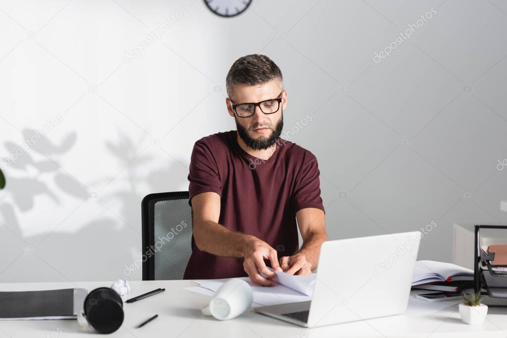 Businessman holding papers near laptop, cup and clumped paper on table 