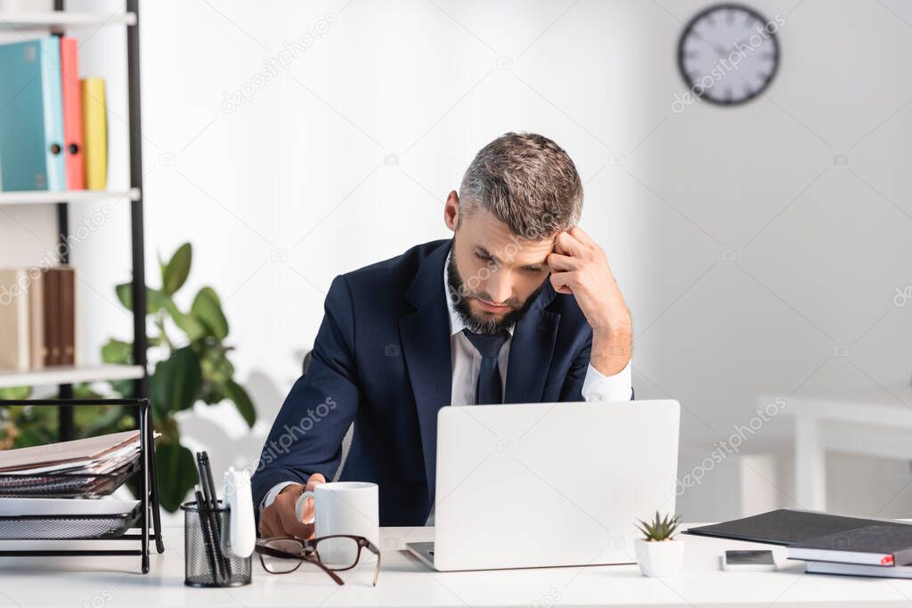 Tired businessman looking at laptop near stationery, cup and eyeglasses on blurred foreground 