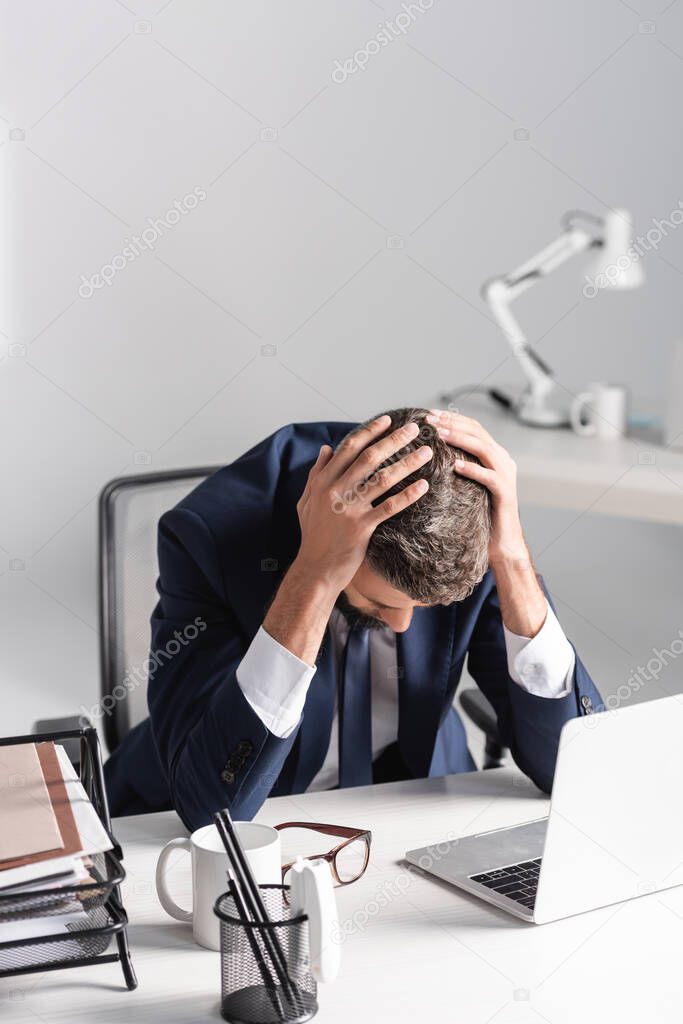 Tired businessman with hands near head sitting beside laptop and stationery on table in office 