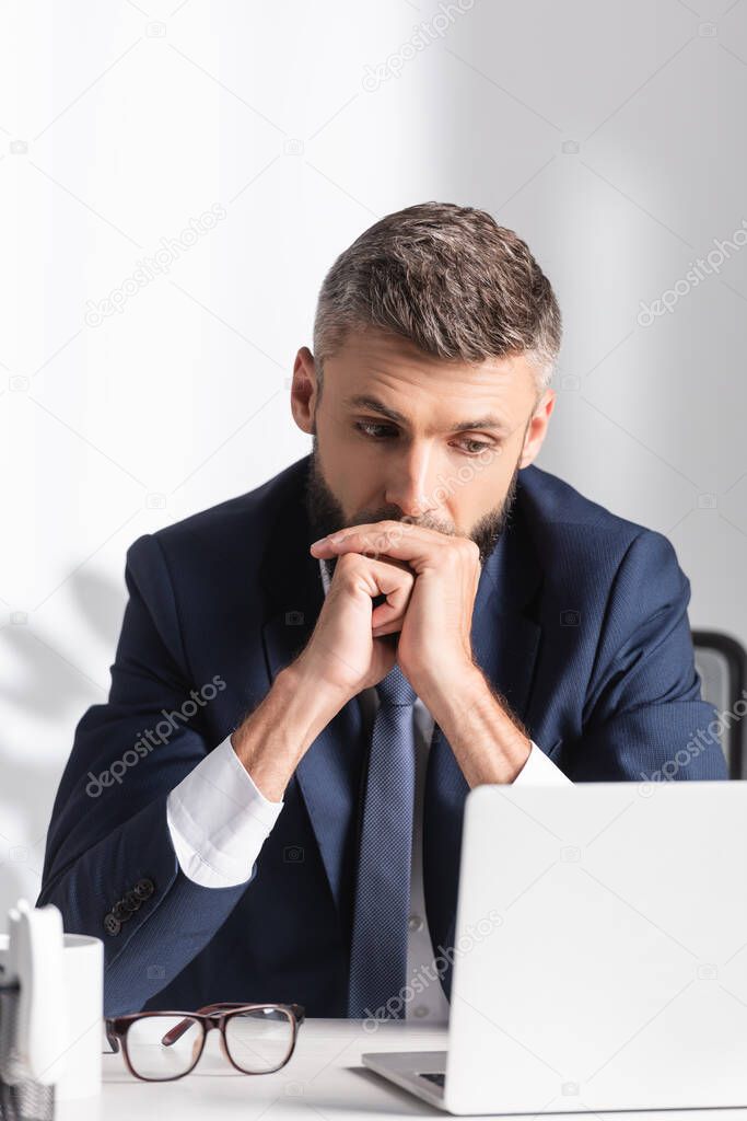 Pensive businessman in suit looking at laptop on blurred foreground in office 