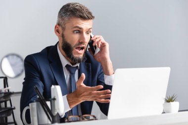 Heyecanlı iş adamı akıllı telefonda konuşuyor ve bulanık önplanda dizüstü bilgisayarı işaret ediyor. 