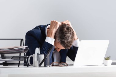 Tired businessman sitting near laptop and documents on blurred foreground in office  clipart