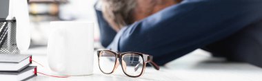 Eyeglasses and cup on table near tired businessman on blurred background in office, banner  clipart