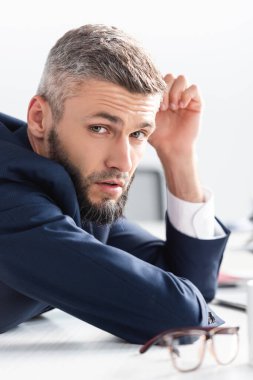 Takım elbiseli bir işadamı masanın önündeki bulanık ön planda gözlüklü kameraya bakıyor. 