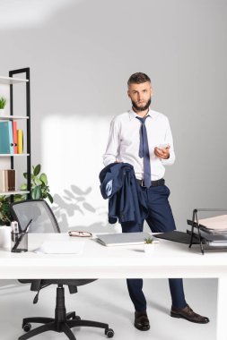 İş adamı gazetelerin yanında akıllı telefon ve ceket tutuyor ve ön plandaki bulanık çalışma masasında kırtasiye malzemesi tutuyor. 