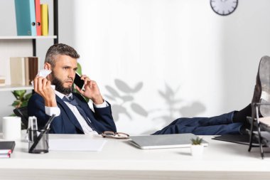 İş adamı bilgisayarın yanında akıllı telefonla konuşurken kağıt yığınını tutuyor ve ön planda bulanık kırtasiye malzemesi var. 
