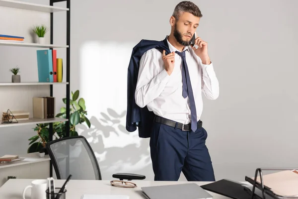 Empresario Sosteniendo Chaqueta Hablando Teléfono Inteligente Cerca Computadora Portátil Papeles —  Fotos de Stock