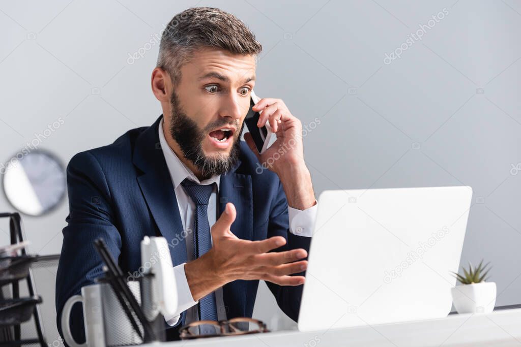 Excited businessman talking on smartphone and pointing at laptop on blurred foreground 