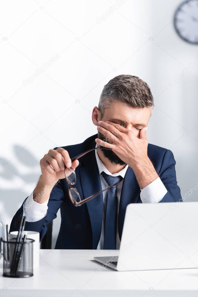 Tired businessman holding eyeglasses and touching eyes near laptop on blurred foreground 