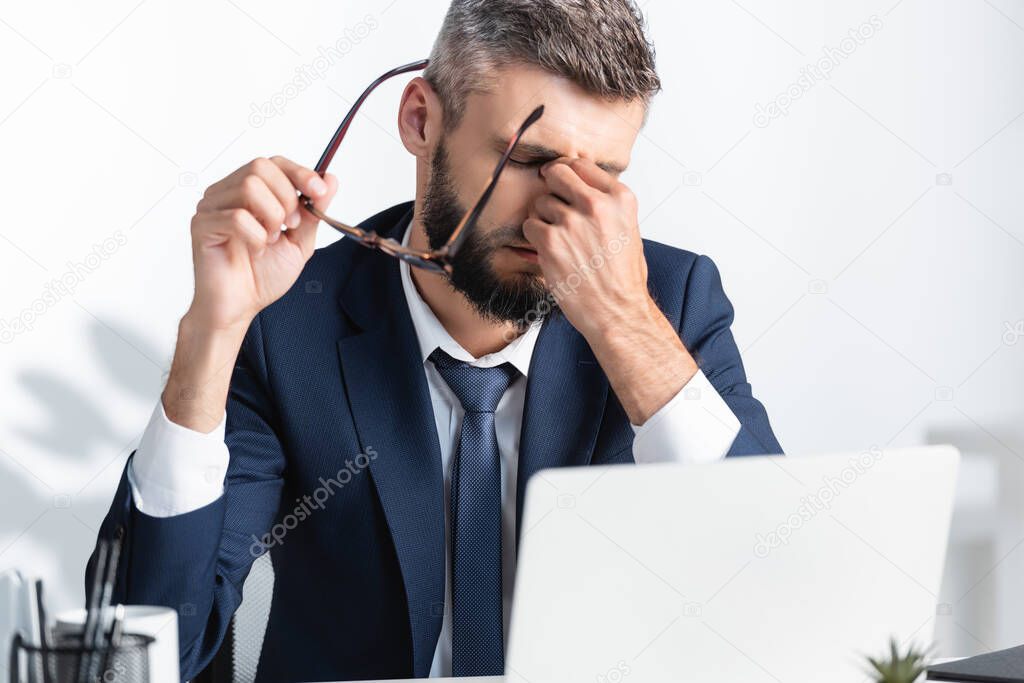 Tired businessman holding eyeglasses while working near laptop on blurred foreground 