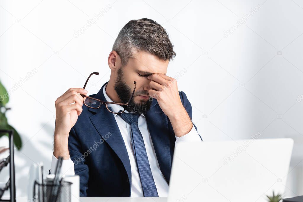 Overworked businessman holding eyeglasses and touching eyes near laptop on blurred foreground 