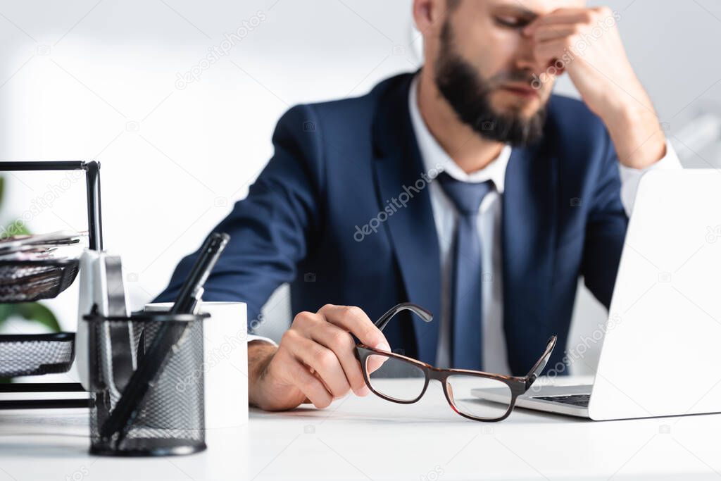 Stationery and cup on table with overworked businessman holding eyeglasses near laptop on blurred background 