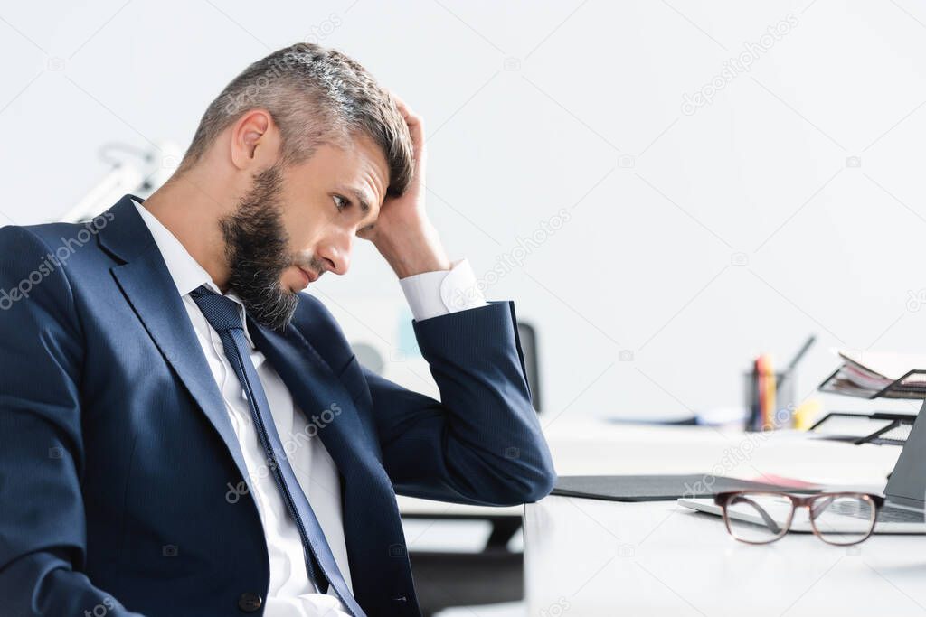 Tired businessman looking at laptop near eyeglasses on blurred foreground on office table 