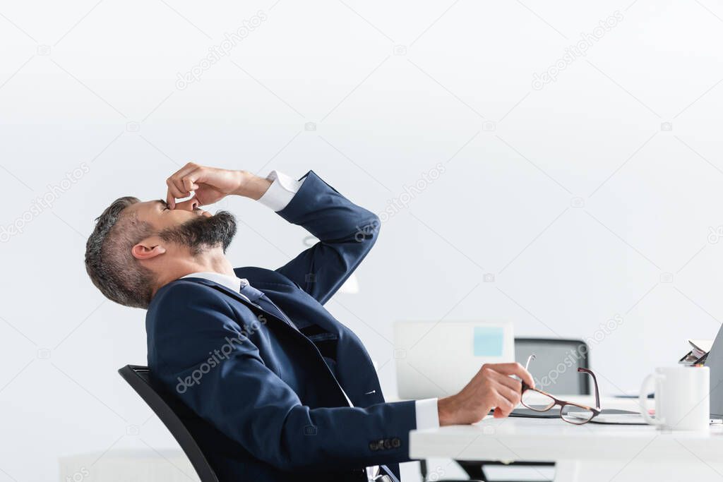 Overworked businessman in formal wear touching eyes while holding eyeglasses in office 