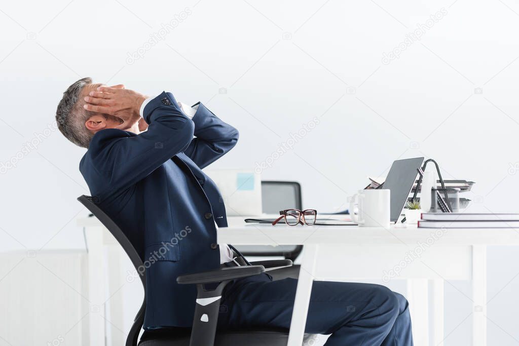 Tired businessman touching face near laptop and eyeglasses on blurred foreground in office  