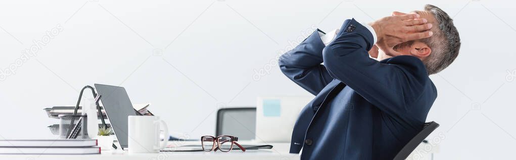 Tired businessman touching face near laptop and eyeglasses on blurred foreground in office, banner