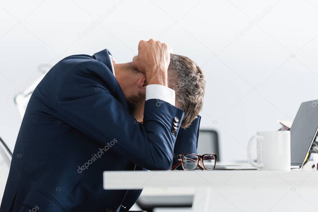 Exhausted businessman in suit touching head while working near laptop, eyeglasses and cup in office 