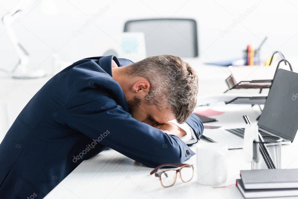 Tired businessman with closed eyes sitting near laptop and stationery on blurred foreground in office 