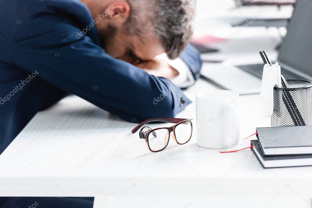 Eyeglasses, cup and stationery on table near overworked businessman sleeping on blurred background 