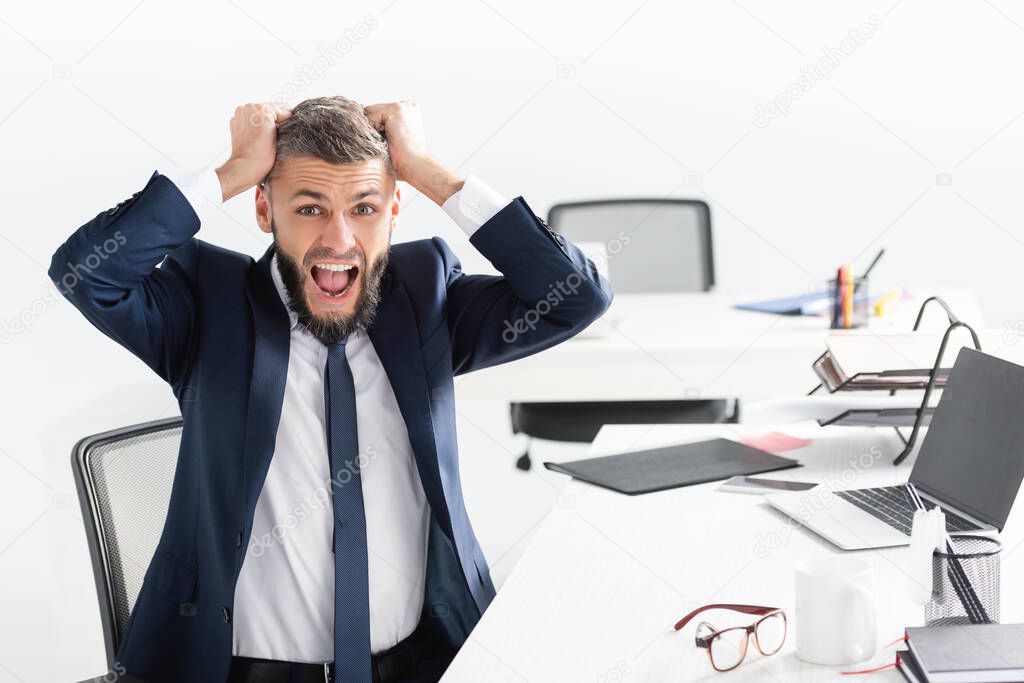 Nervous businessman screaming near eyeglasses and laptop on blurred foreground in office 
